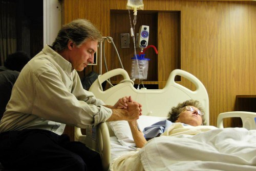 man-sitting-with-older-woman-in-hospital-bed-holding-her-hand